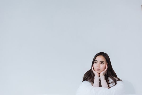 A thoughtful Asian woman gazes pensively with hands on chin in a minimalist studio.