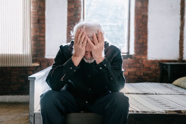 An elderly man sitting on a bed indoors, expressing grief and loss with his head in his hands.