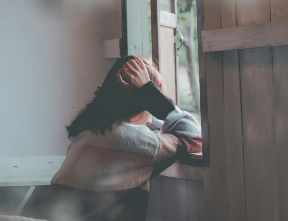 A woman sits indoors gazing thoughtfully out of a window, evoking emotions of loneliness and reflection.