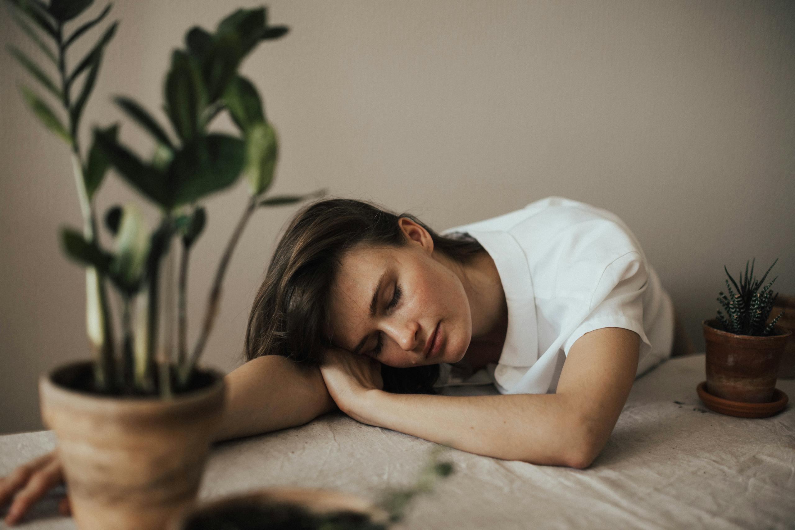 A woman peacefully resting her head on a table surrounded by indoor plants.