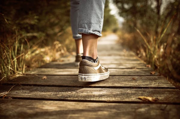 Person wearing sneakers walking on a wooden path in a sunlit park.