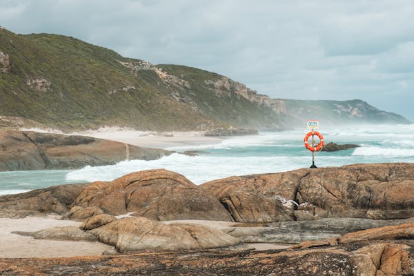 Scenery of foamy azure sea waves crashing on rocky rough seashores with lifebuoy on pole under cloudy gloomy sky