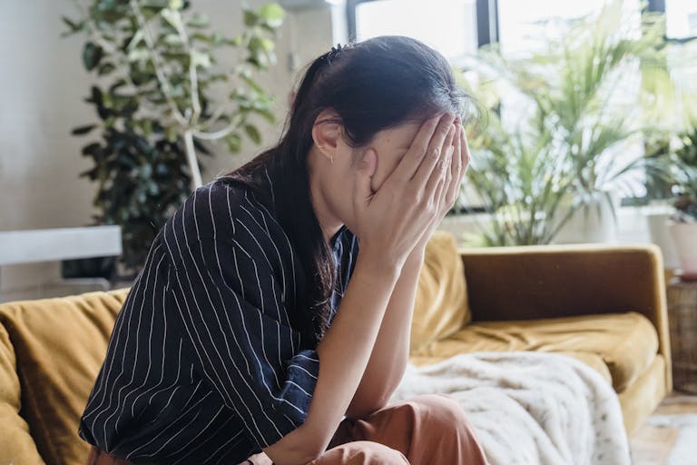 Woman covering face with hands, looking frustrated while sitting on a sofa in a modern living room.