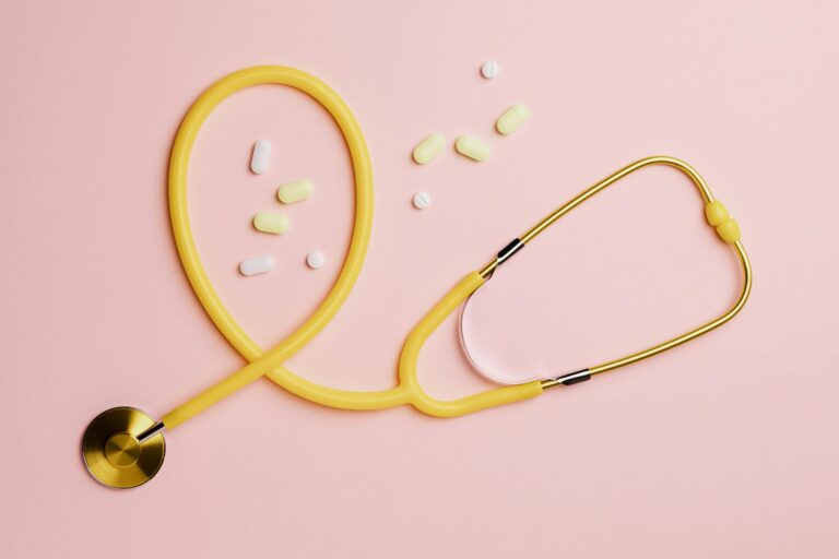 Flat lay of a yellow stethoscope and assorted pills on a pastel pink background.