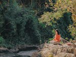 Woman meditating by a serene riverside surrounded by lush greenery on a sunny day.