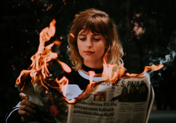 A young woman reading a newspaper on fire outdoors, with flames lighting up the scene.