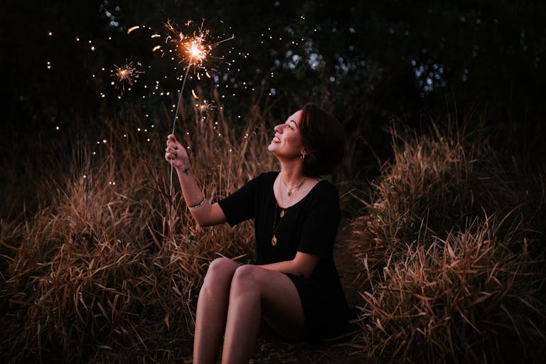 A smiling woman enjoying sparklers outdoors in a grassy field at night, expressing pure joy.