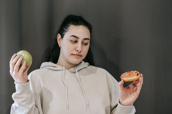 A woman in a hoodie contemplates a healthy apple versus a sugary doughnut.