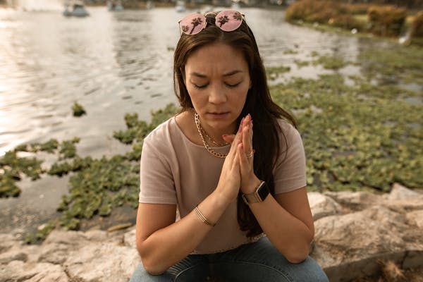 A woman sits by a lake, deep in thought with hands clasped in contemplation.