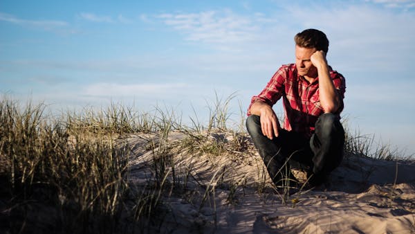 Man in a red plaid shirt sitting thoughtfully on a sandy dune in Australia, surrounded by grass.