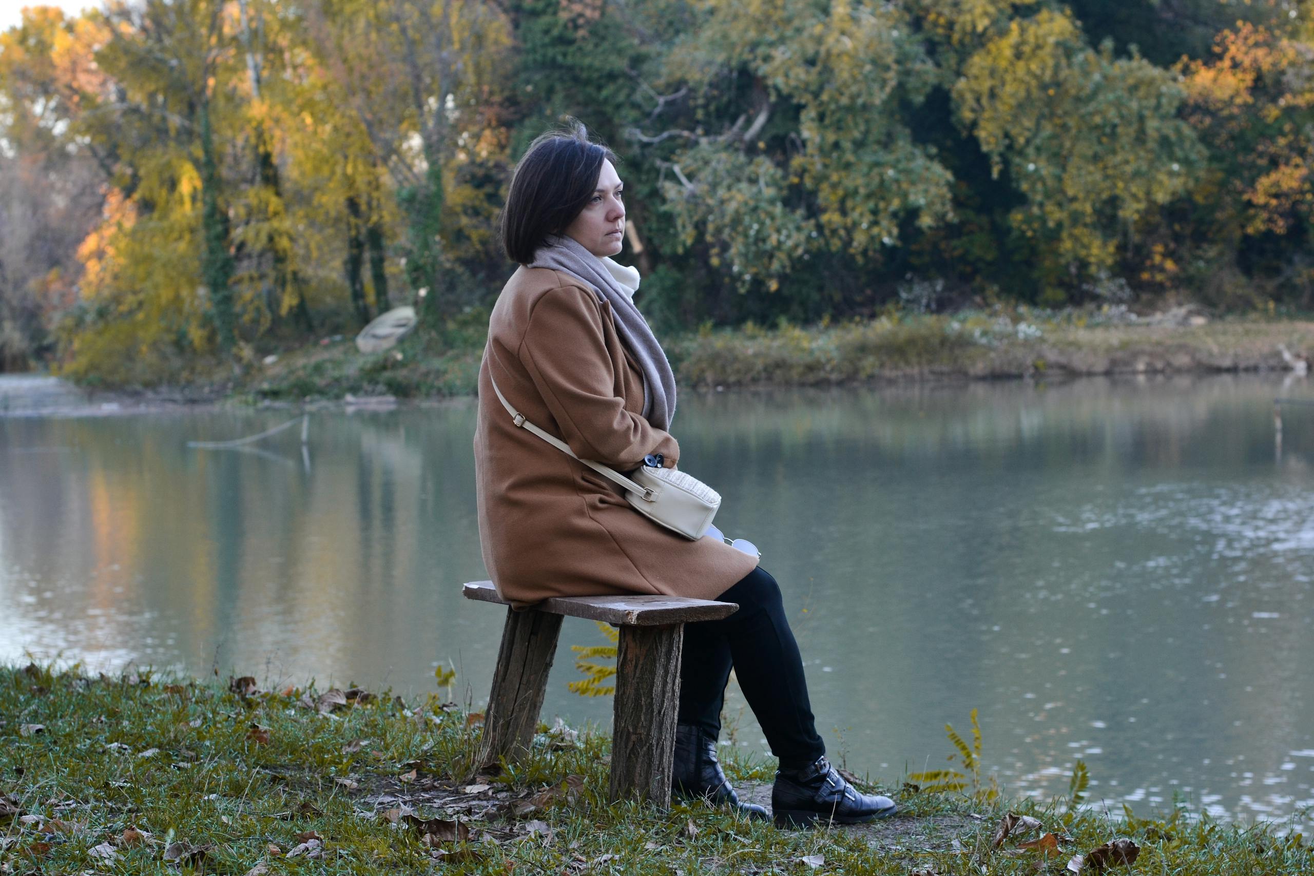 Woman in a brown coat sitting by a tranquil lakeside in autumn, lost in thought.