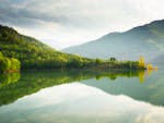 A tranquil lake reflecting lush green mountains and trees under a cloudy sky.