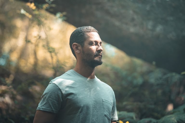 A man with short hair and beard stands calmly outdoors, immersed in nature.